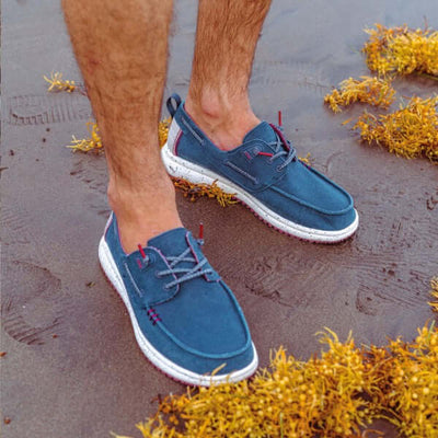 Person wearing Byron Ultralight Boat Shoes with blue canvas and white sole, standing on sand near seaweed.