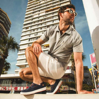 Man wearing Maui Hi-Grip Boat Shoes with blue uppers and red soles, posing in urban setting beside palm trees and tall building.