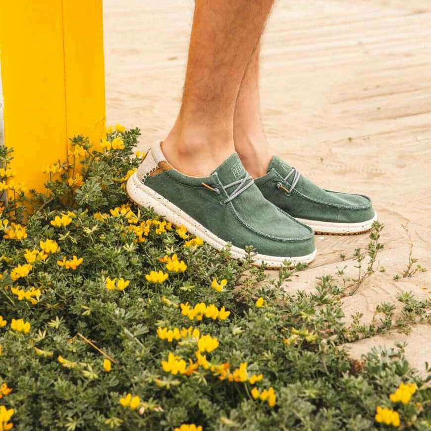 Green Nias Ultralight Wally boat shoes with easy-on elastic laces, worn outdoors next to yellow flowers on a wooden deck.